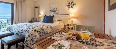 A hotel room with a bed, a dining table with breakfast, a view of the ocean, and a ship decoration on the wall.