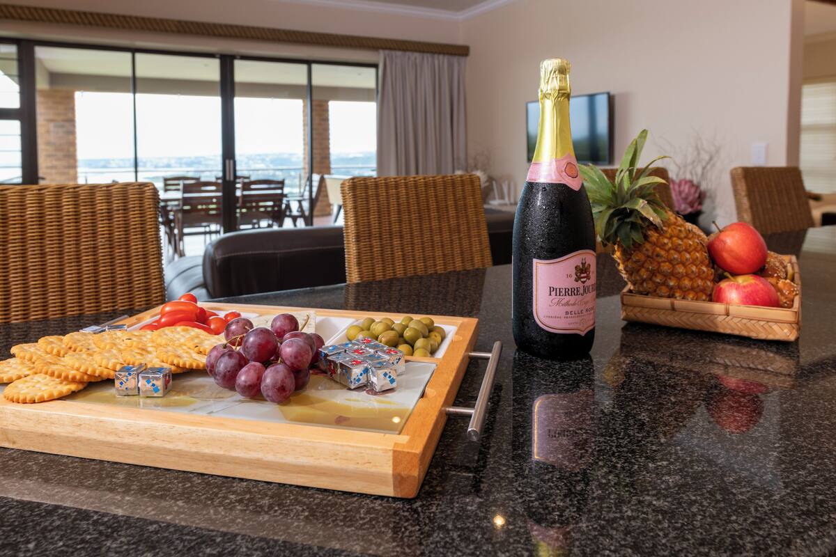 A table with a bottle of champagne, a fruit basket, and a cheese board with crackers and grapes.