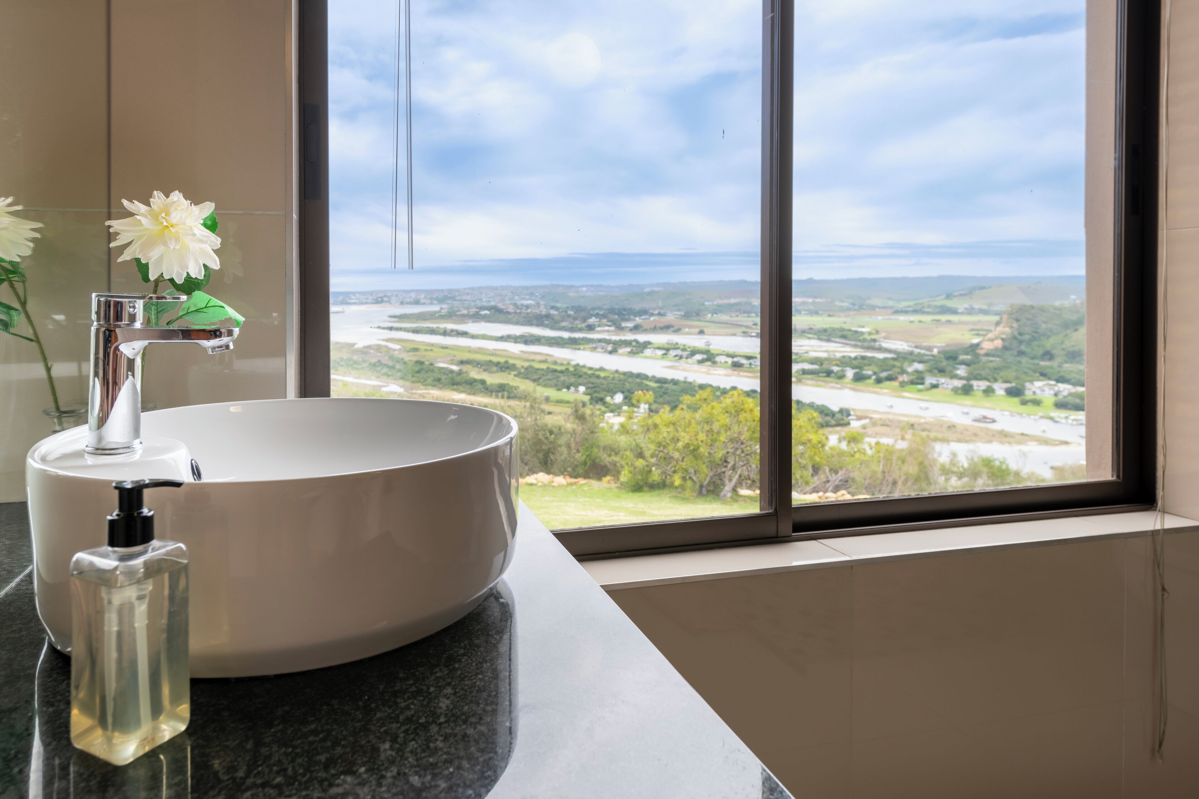 A bathroom with a large window, a white sink, and a clear glass bottle.