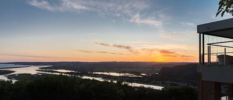 Vista al atardecer desde un edificio moderno con balcón de vidrio que da a un río y al paisaje circundante.