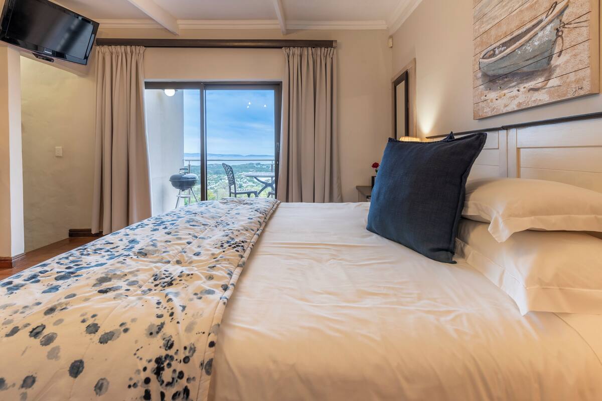 A hotel room with a bed, a flat-screen TV, a painting on the wall, and a balcony with a view.
