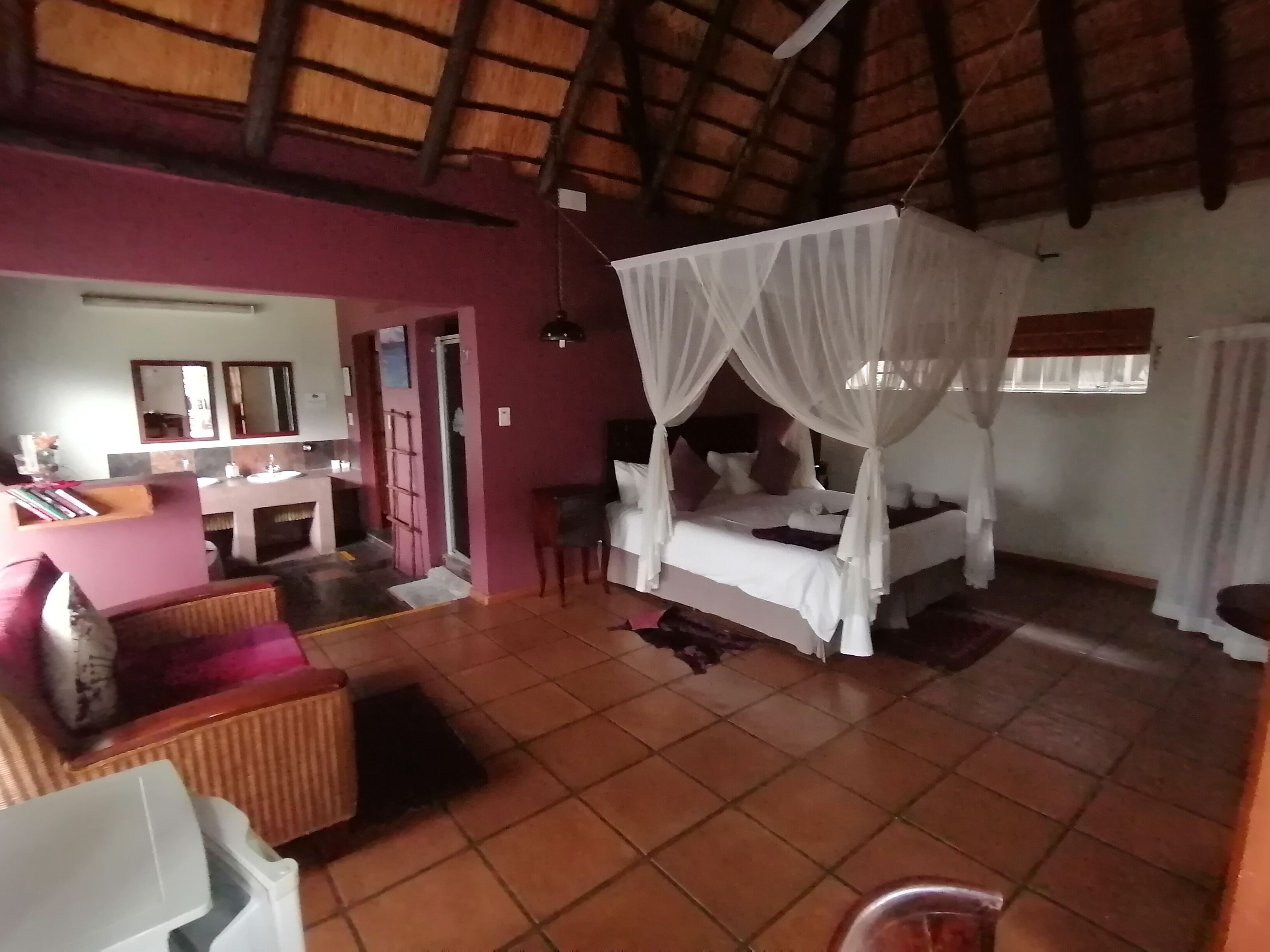 A room with a bed under a canopy, a bathroom with a sink and mirror, and a chair.