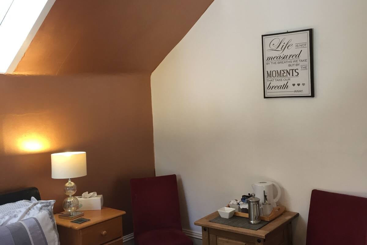 A bedroom with a bed, a nightstand, a lamp, a table with a kettle, two chairs, and a wall-mounted quote.