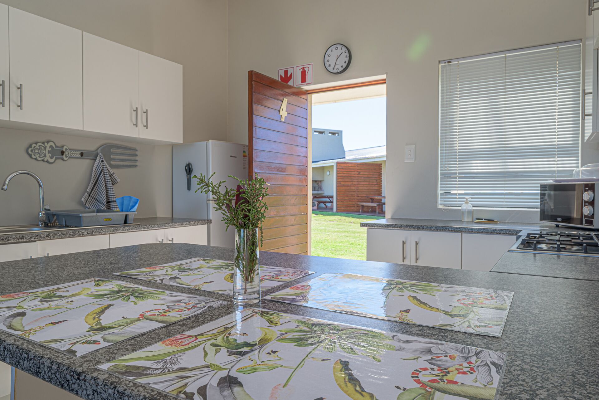 A modern kitchen with white cabinets, a stainless steel sink, a clock, and a window with blinds.
