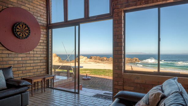 A room with large windows overlooking a beach, a dartboard, and a leather sofa. - Fynbos Golf & Country Estate (Eersterivierstrand)