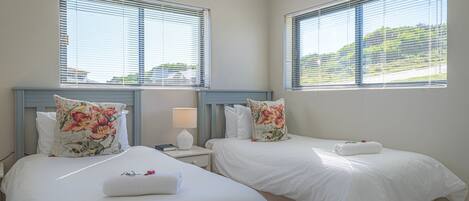 A hotel room with two beds, each with white bedding and floral pillows. There is a nightstand with a lamp and a book between the beds.