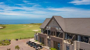 A modern building with a golf course in the background, a covered parking area with golf carts, and a view of the ocean. - Fynbos Golf & Country Estate (Eersterivierstrand)