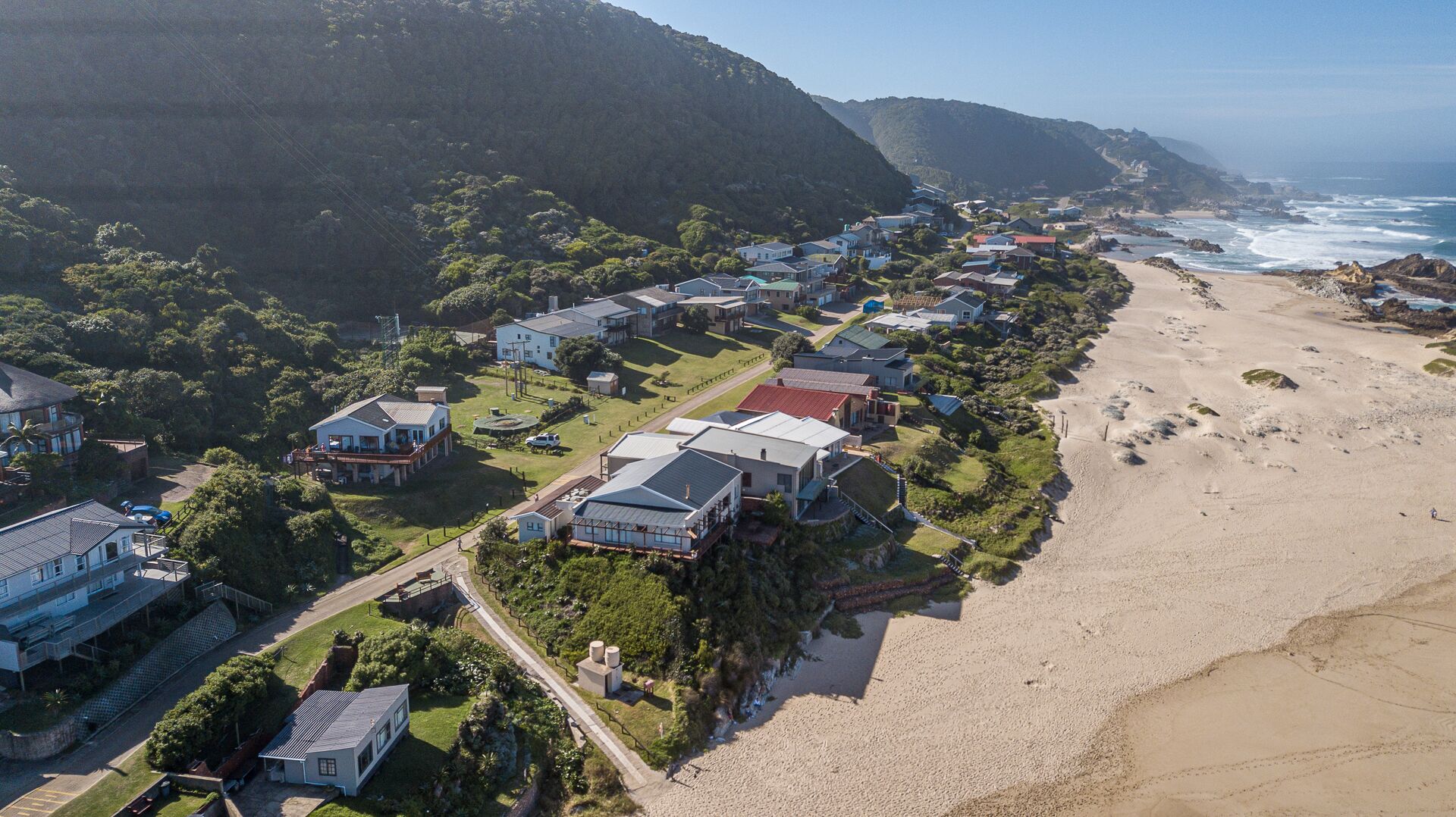 An aerial view of a coastal residential area with houses, a beach, and a mountainous backdrop.