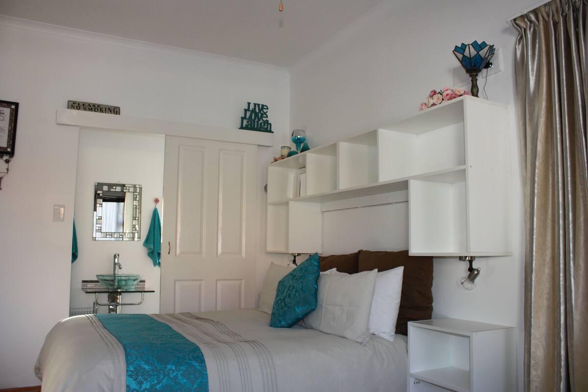 A bedroom with a bed, a nightstand, a mirror, a bathroom vanity, and a shelf with decorative items.
