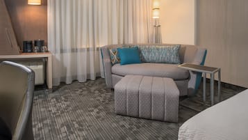 A hotel room with a sofa, ottoman, a small table, a desk with a lamp, and a bed.