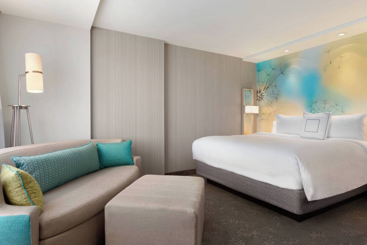 A modern hotel room with a bed, a sofa, a standing lamp, and a textured wall.