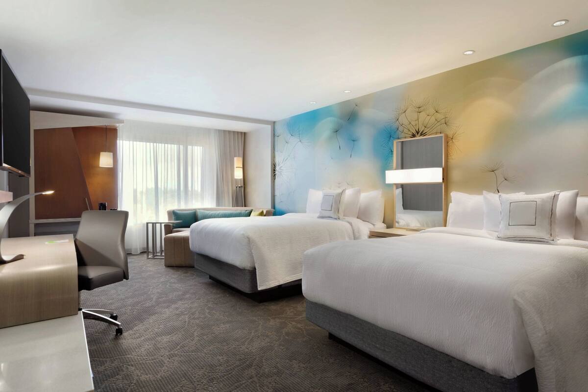 A hotel room with two beds, a desk, and a chair. There is a large window with curtains, a wall-mounted TV, and a decorative wall piece.