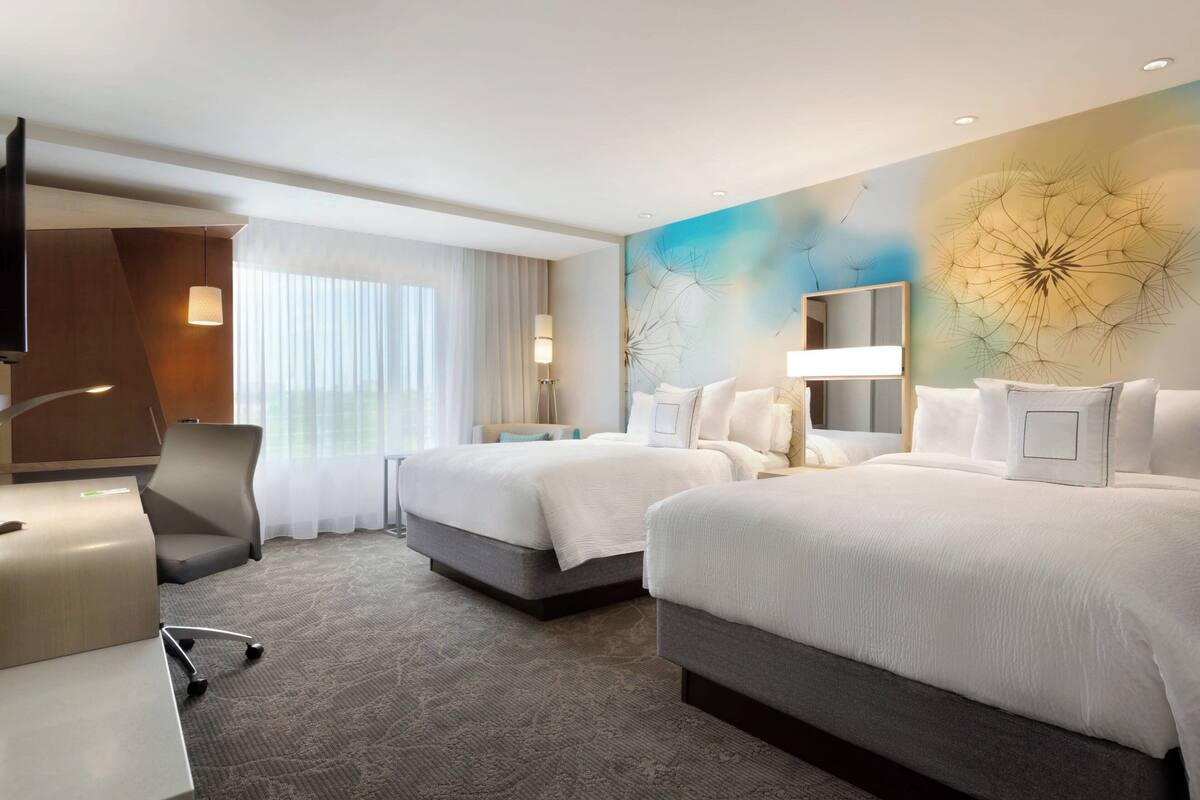 A hotel room with two beds, a desk, a chair, a large window with curtains, and a wall with a dandelion design.
