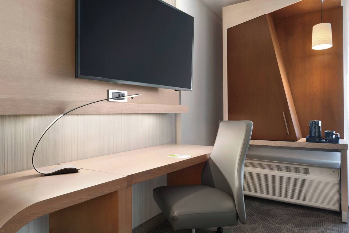 A modern hotel room with a flat-screen TV mounted on the wall, a wooden desk with a chair, a coffee machine, and a heater.
