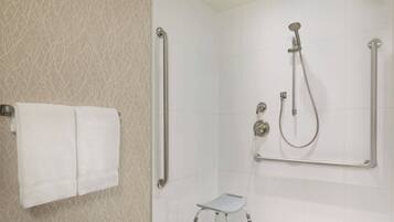Un baño con pared de azulejos blancos, una toalla blanca colgada en la pared, una ducha con cabezal de ducha manual y barras de apoyo, y un taburete pequeño.