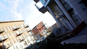 A multi-story residential building with balconies, a paved ground, and a clear sky.