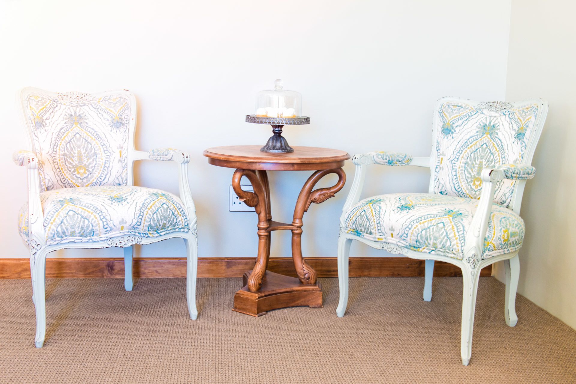 Two white chairs with patterned upholstery, a small round wooden table with a glass top, and a decorative lamp.