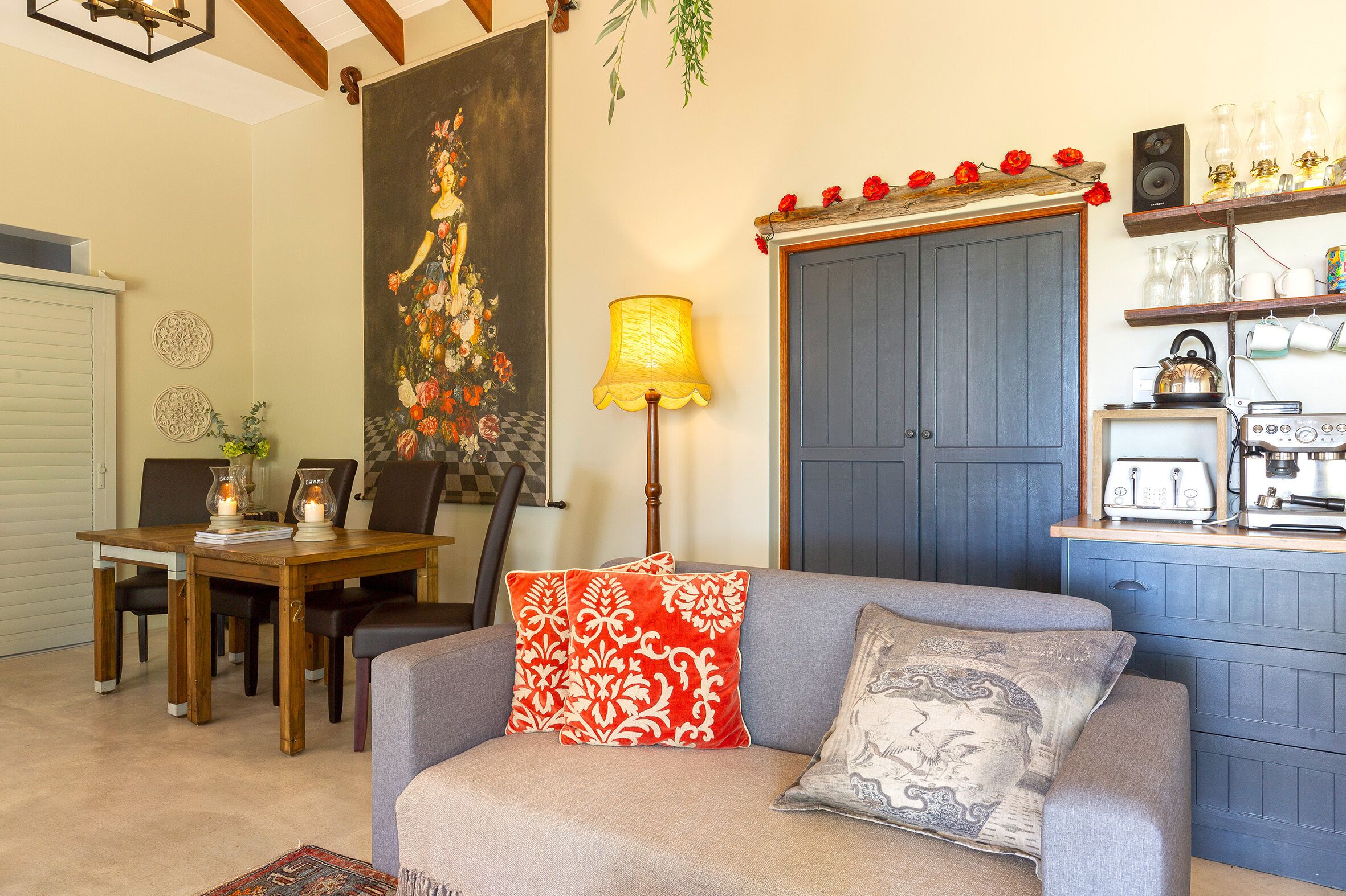 A living room with a sofa, a dining table with chairs, a lamp, a painting, and a kitchen area with appliances.