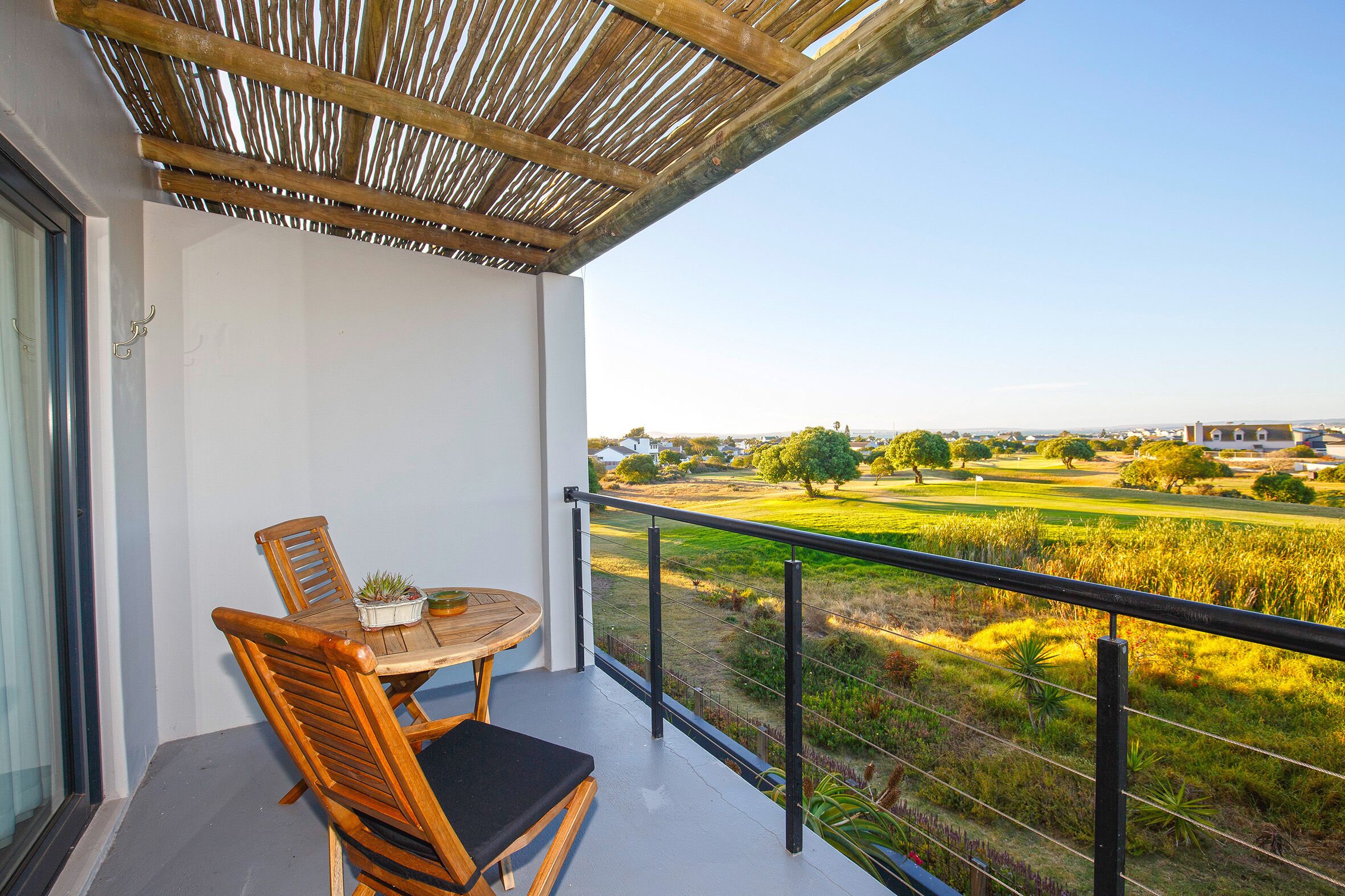 A balcony with a wooden table and chairs, a view of a green landscape with fields and trees, and a clear sky.