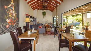 A dining area with wooden tables and chairs, a kitchen in the background, and a large window providing a view of the outdoors.