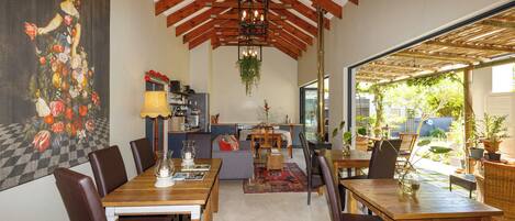 A dining area with wooden tables and chairs, a kitchen in the background, and a large window providing a view of the outdoors.
