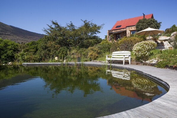 A serene outdoor pool area with a wooden deck, a bench, and a stone building with a red roof in the background.