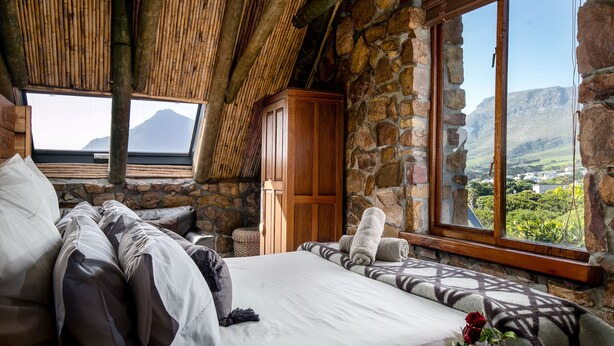 A bedroom with a large bed, stone walls, a wooden wardrobe, and a window with a mountain view. - Leopardstone Hill (Cape Town)