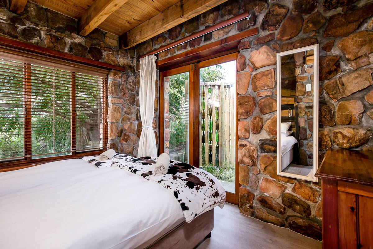 A bedroom with a stone wall, wooden ceiling, a large bed with a cowhide blanket, and a view through a window with blinds.