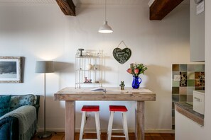 A kitchen with a wooden table, two red stools, a hanging light, a heart-shaped sign, a vase with flowers, and a tiled backsplash.