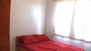 A small, single-bed room with a wooden frame, a red bedspread, and a window with a white curtain. - Ximple Appartementen (Paramaribo)
