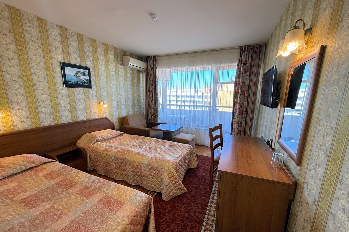 Habitación de hotel con dos camas, un escritorio, una silla, un televisor y una ventana con cortinas.