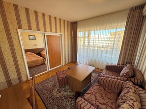 Un salón con un conjunto de sofá, una mesa de centro de madera, una alfombra estampada, un puerta corredera y vistas a través de cortinas traslúcidas.