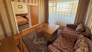 En stue med en sofasæt, et træsofabord, et mønstret tæppe, en skydedør og udsigt gennem lette gardiner.