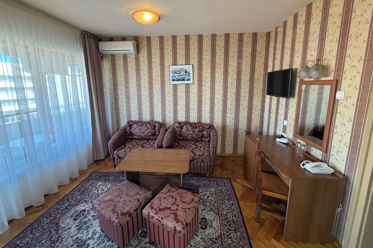 Una habitación con papel tapiz a rayas, un conjunto de sofá, una mesita, otomanas, un televisor y un espejo.