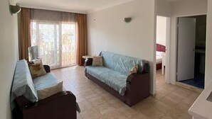 A spacious living room with a sofa set, a television, and a large window with curtains. - Basil's Apart Hotel (Marmaris)