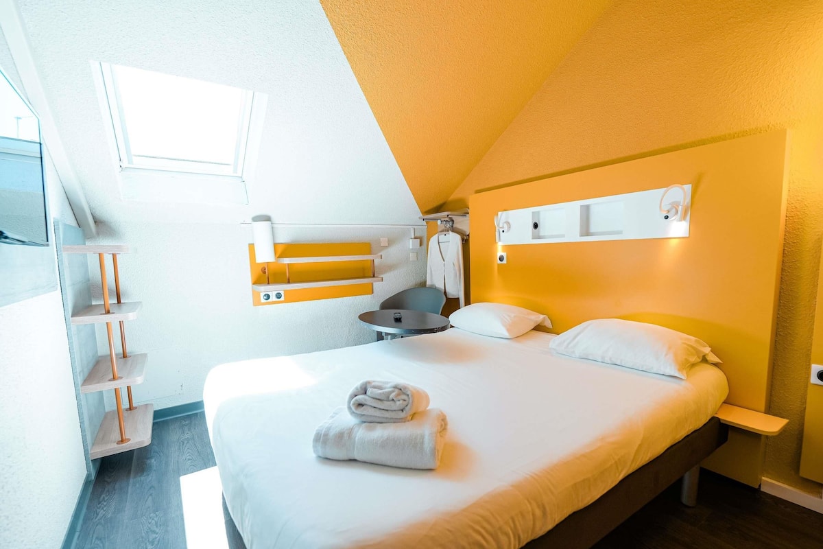 A modern hotel room with a bed, a small table, a chair, and a bathroom area with a towel rack.