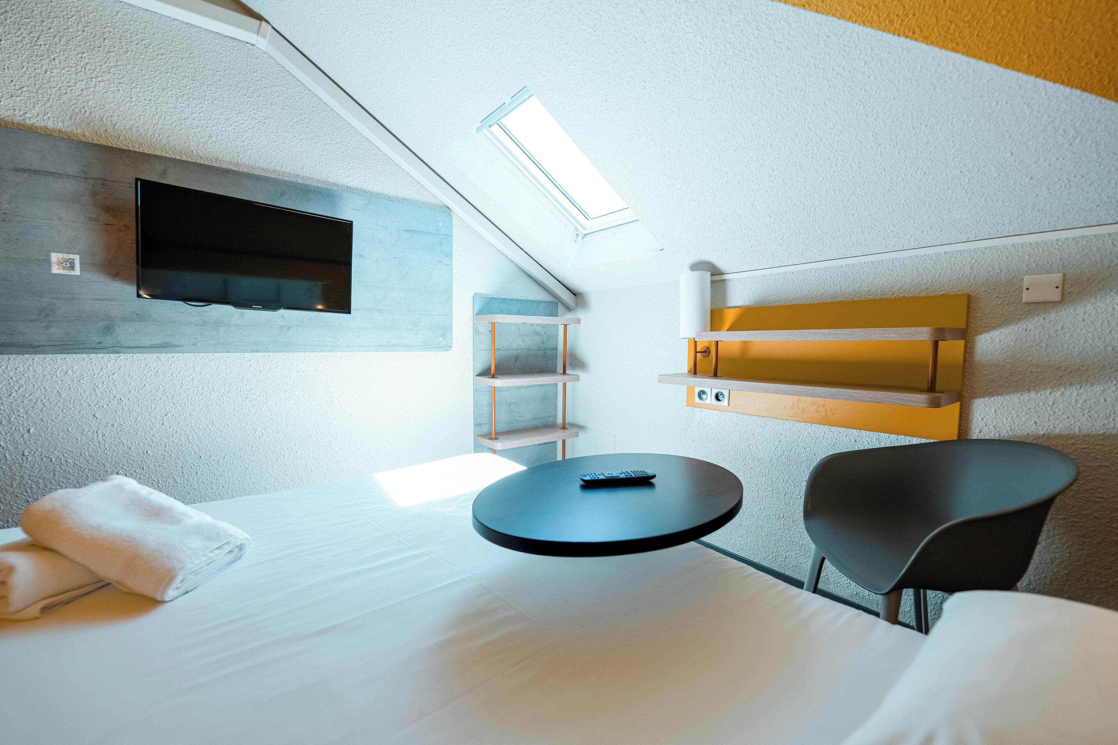 A modern hotel room with a bed, a round table, a chair, a TV mounted on the wall, and a skylight.
