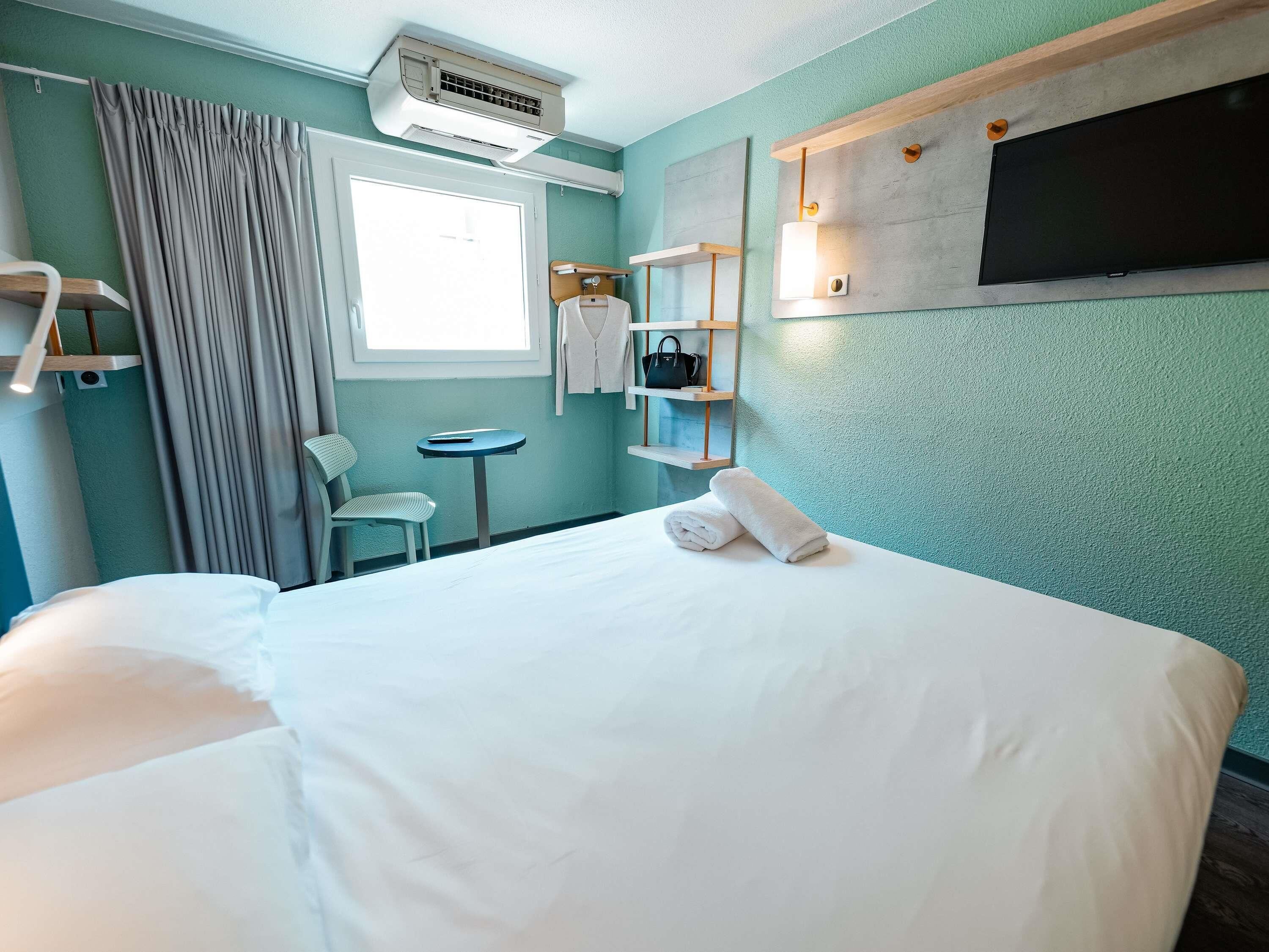 A hotel room with a bed, a chair, a small table, a TV, and a wall-mounted shelf with a lamp.