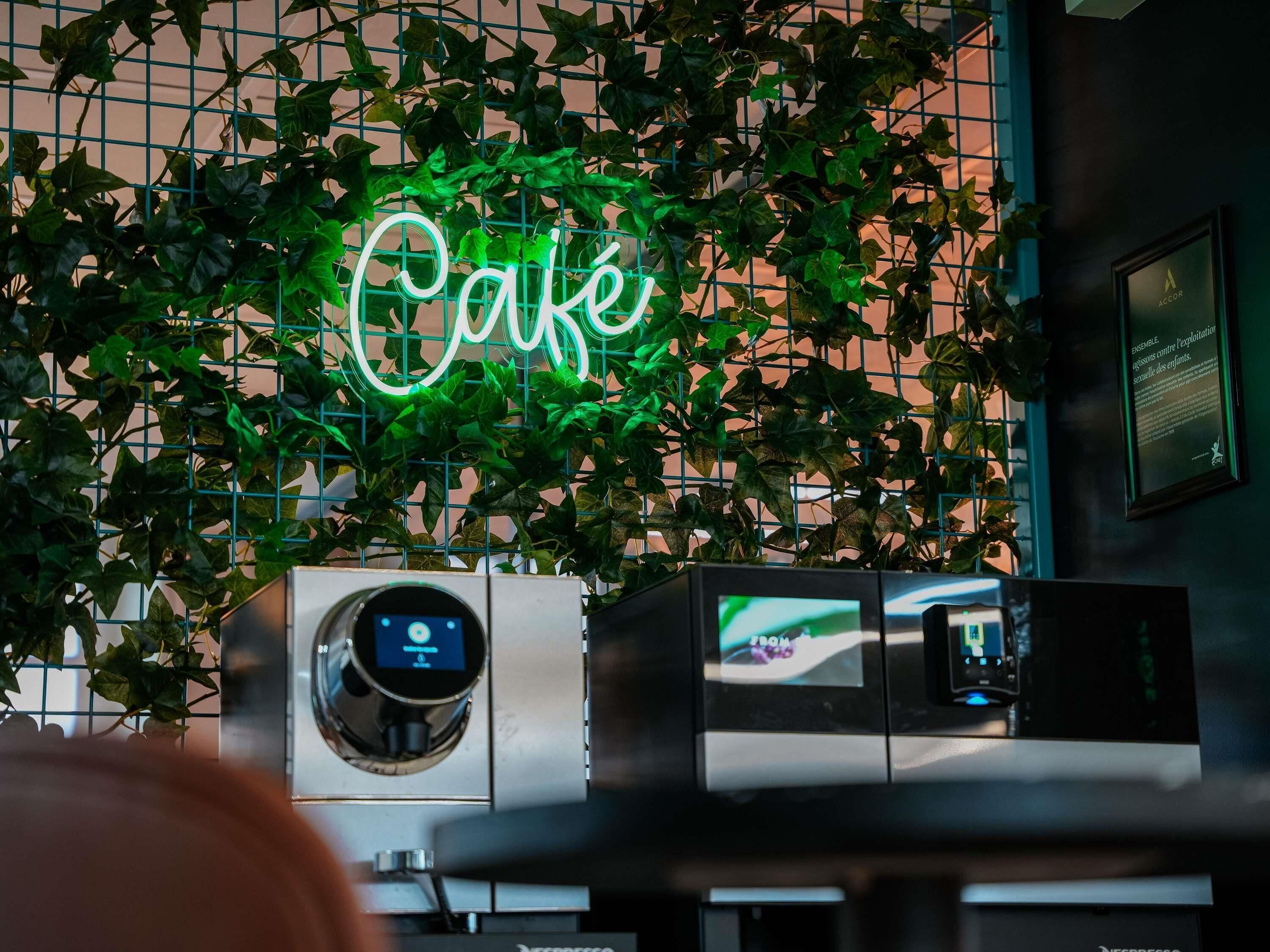 A neon sign reading 'Café' above a coffee machine, with a grid of green plants in the background.