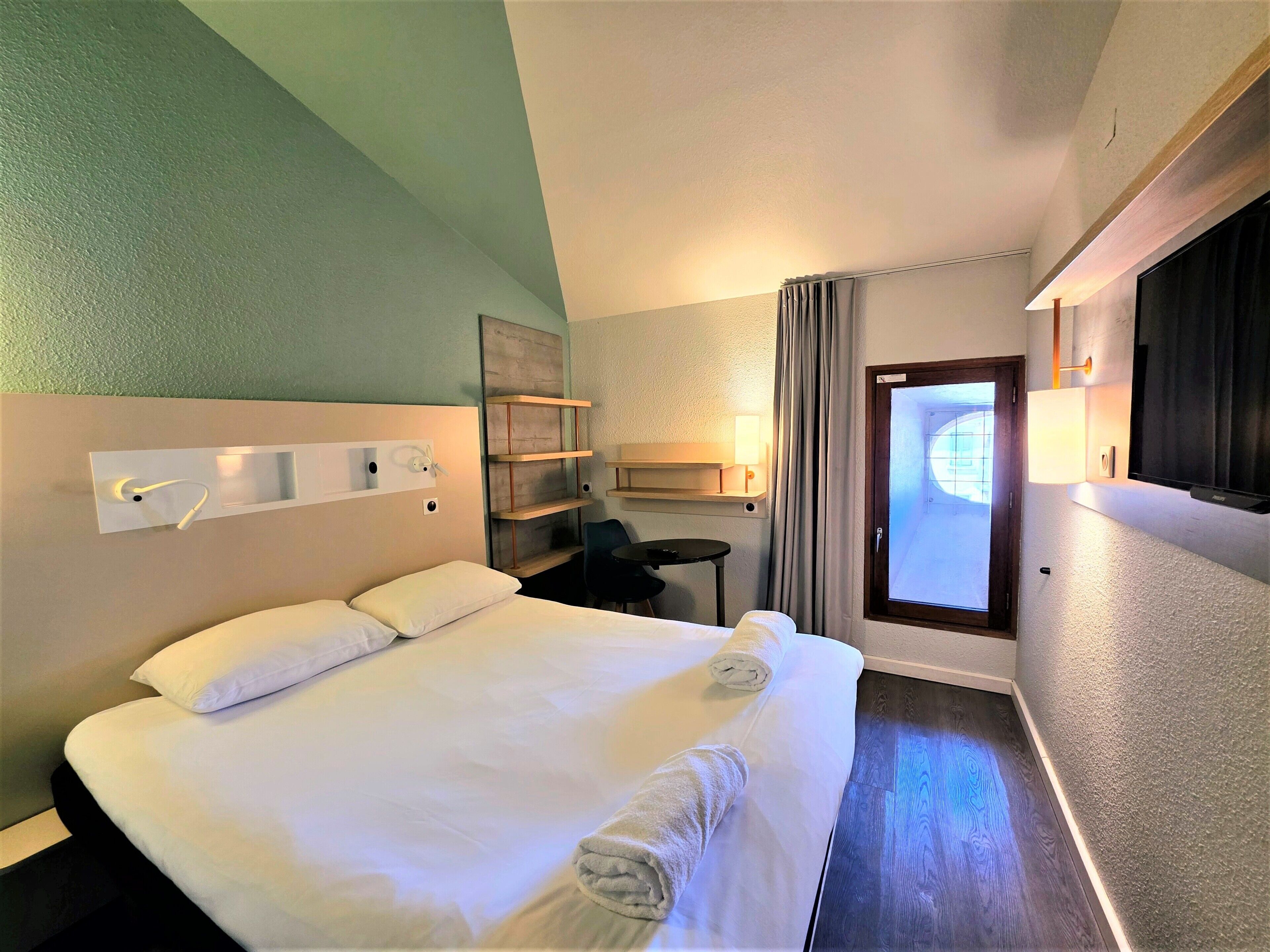 A hotel room with a bed, a desk, a chair, a TV, and a window.