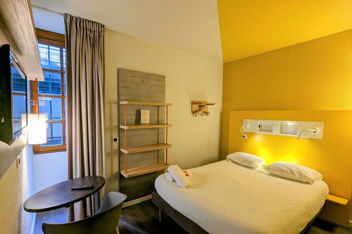A hotel room with a bed, a desk, a chair, a shelf, and a window with curtains.