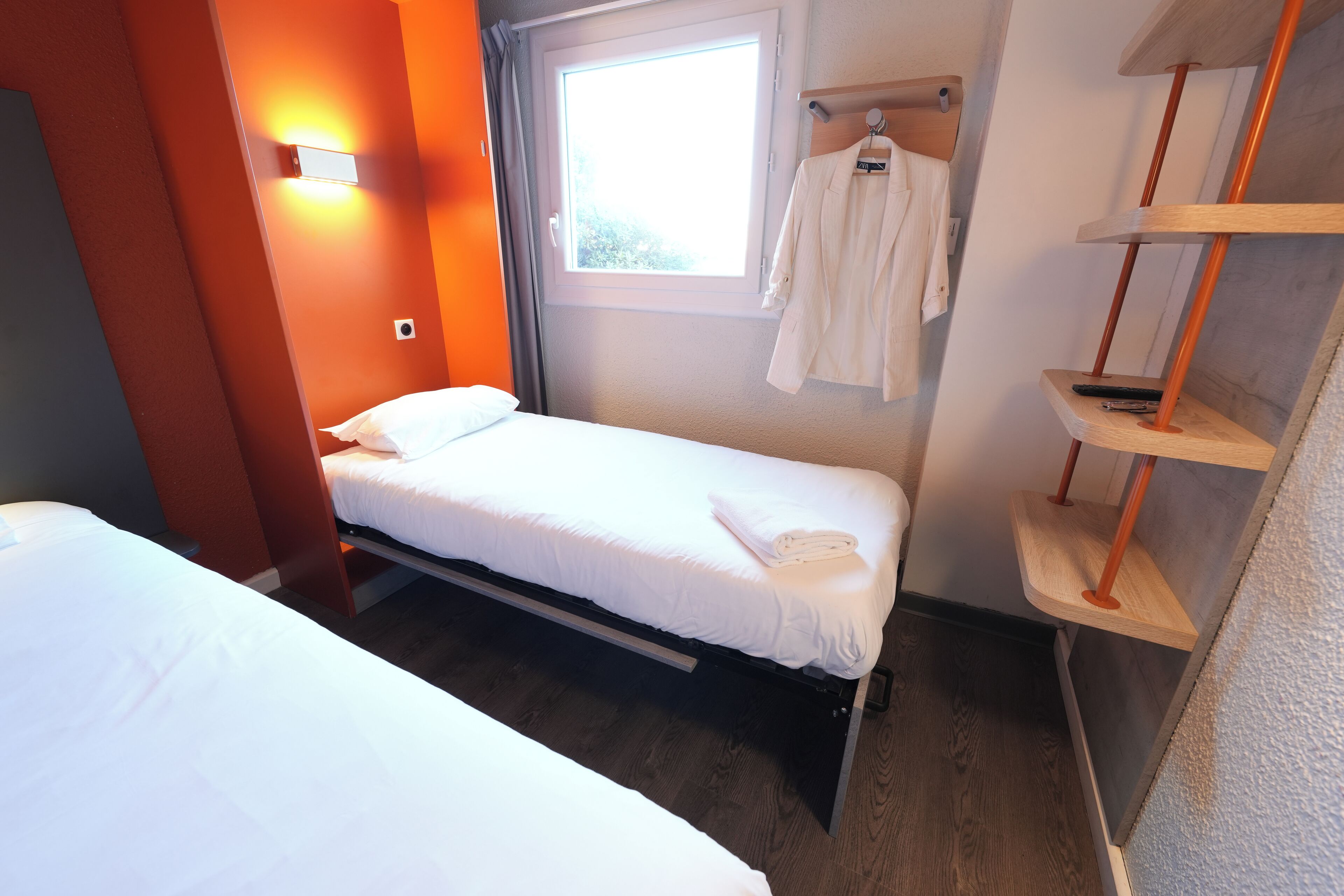 Ein Einzelbett mit weißer Bettwäsche in einem Zimmer mit orangefarbenen Wänden, einem Fenster und einem Holzbodenregal mit einem Handtuch und einer Fernbedienung.