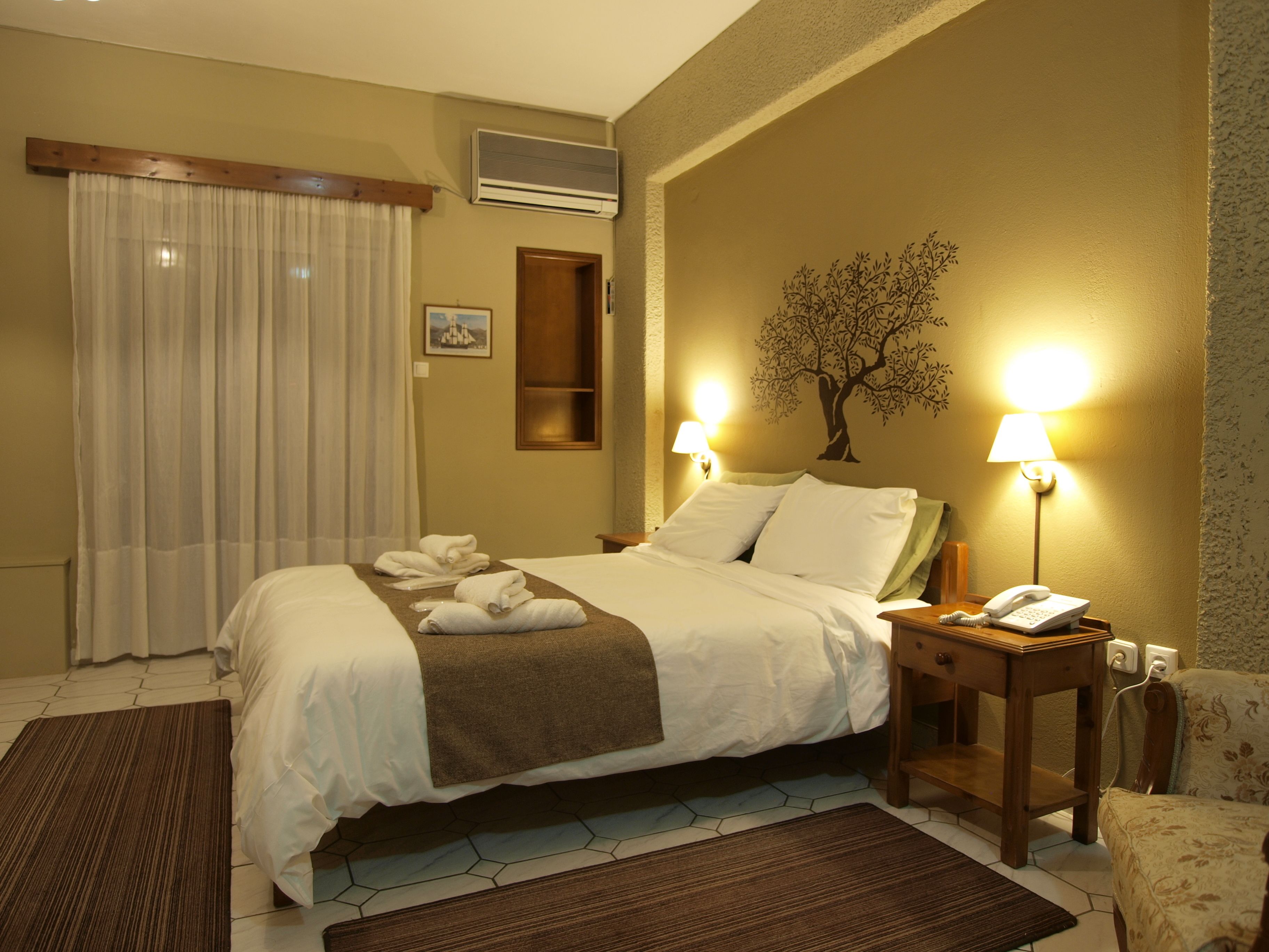 A hotel room with a bed, two bedside lamps, a telephone, a wall-mounted air conditioner, and a window with curtains.