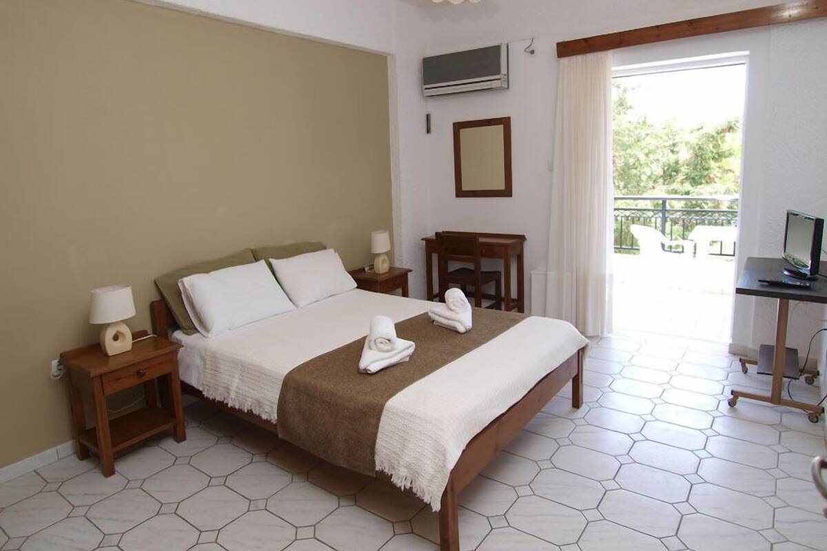 A hotel room with a large bed, a desk with a computer, a TV, and a balcony with outdoor seating.