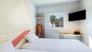 Quarto de hotel com cama, escrivaninha com TV de tela plana, janela com vista para palmeiras e um cabideiro com um casaco.
