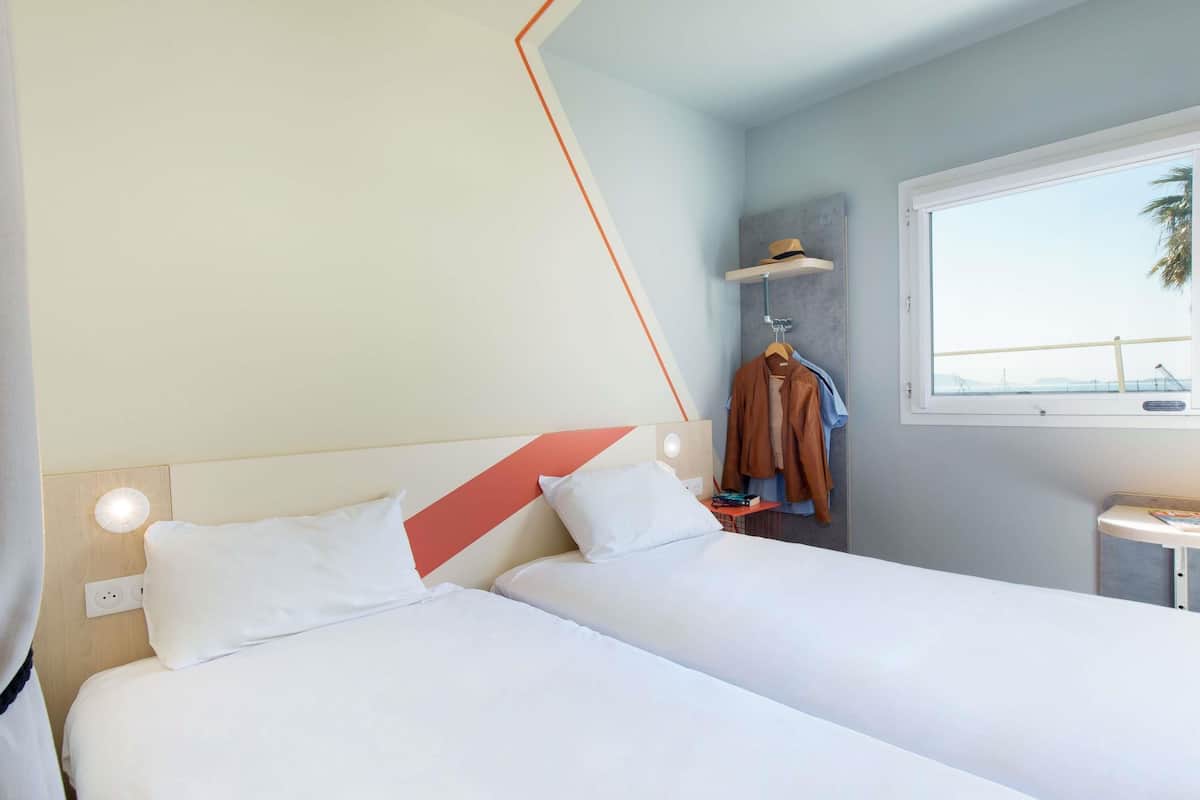 Une chambre d’hôtel avec deux lits, une fenêtre donnant sur un beau paysage, un porte-manteau avec des vêtements et une petite table sur laquelle se trouve un livre.