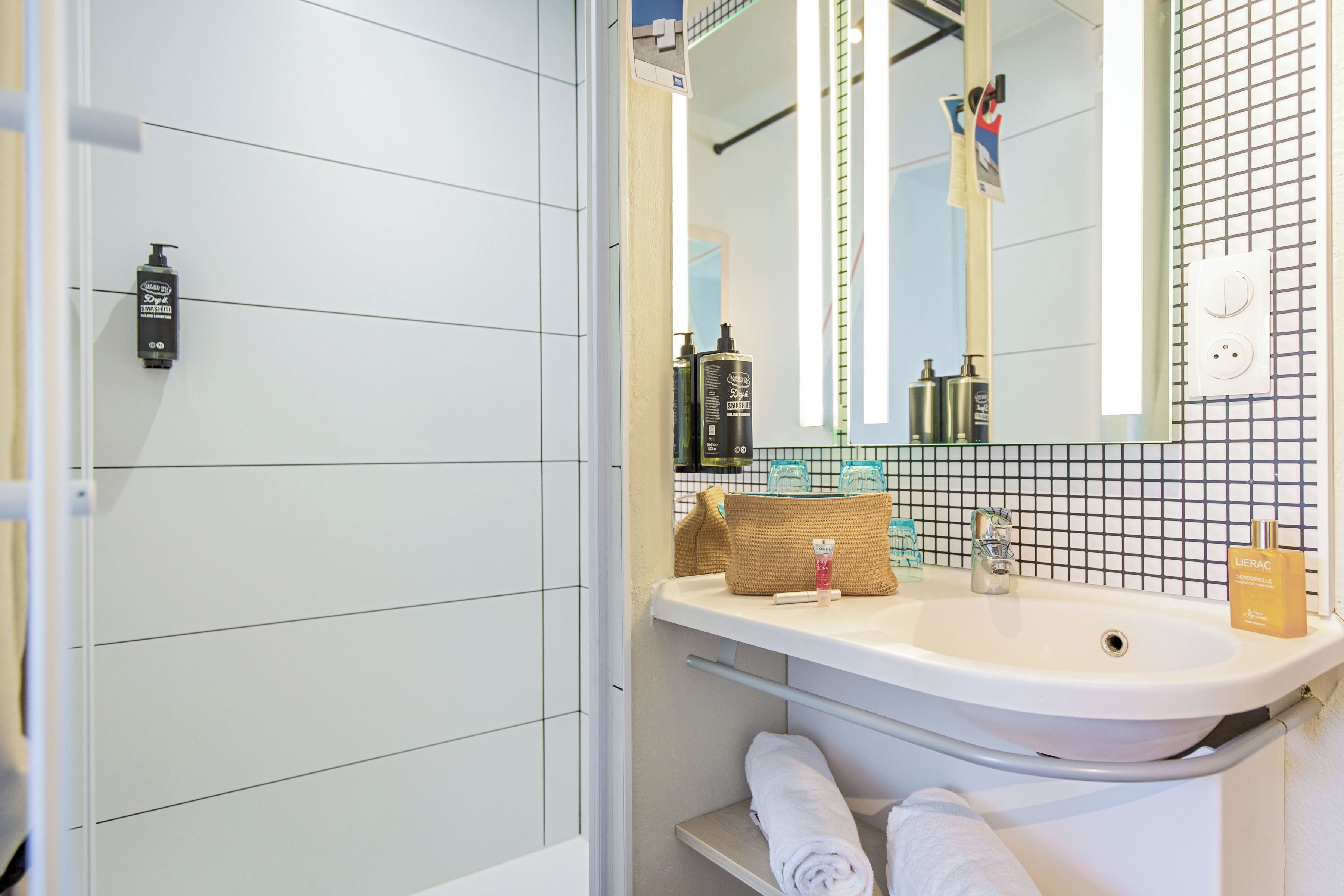 A bathroom with a white sink, mirror, and tiled walls. There are toiletries on the sink and a towel rack below.