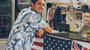 A person leaning on a counter with an American flag backdrop, a store sign, and shelves with products. - World Hostel (Florianópolis)