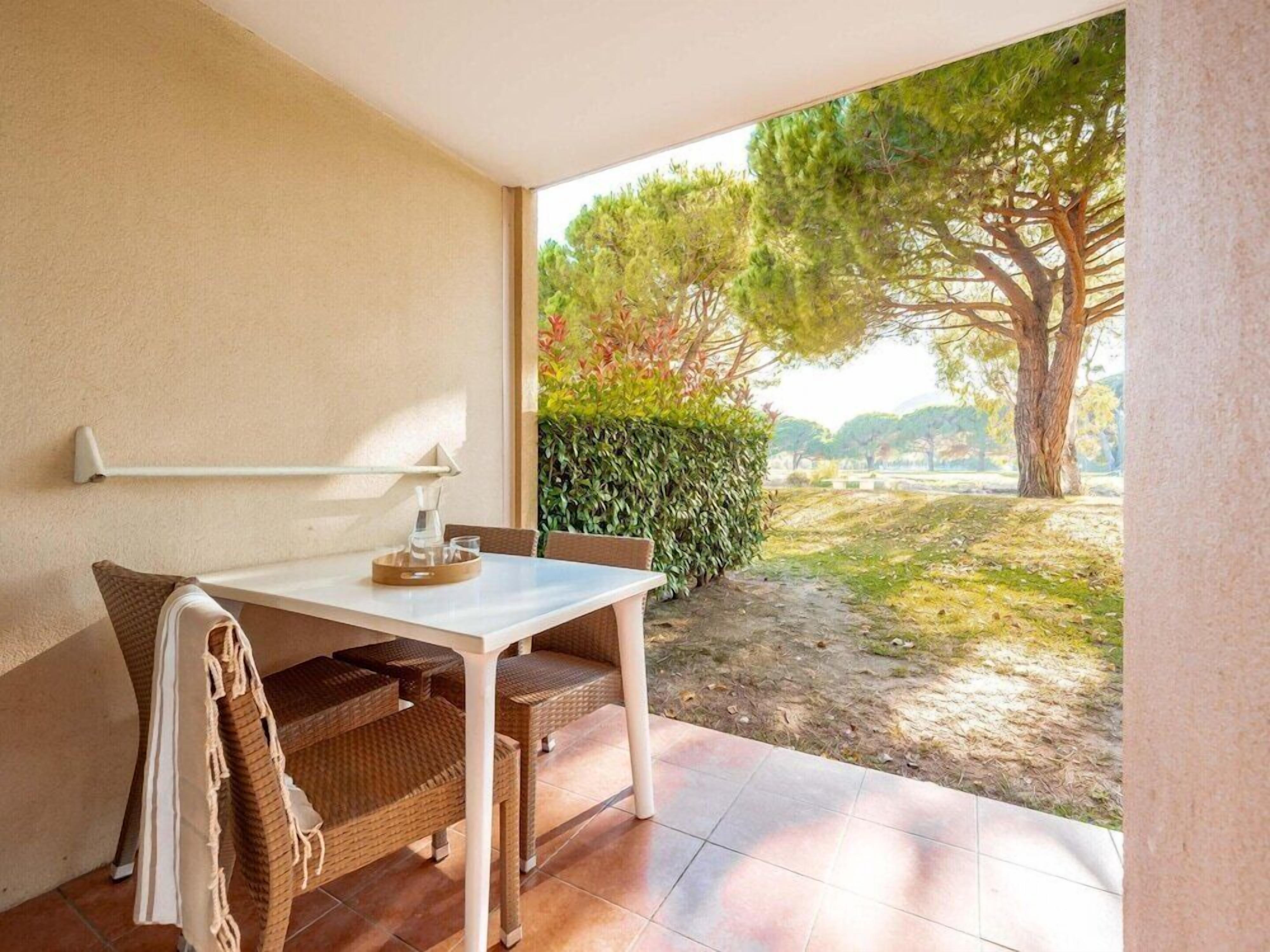 Une petite terrasse avec une table et des chaises, un mur blanc et une vue sur un jardin avec des arbres et une haie.
