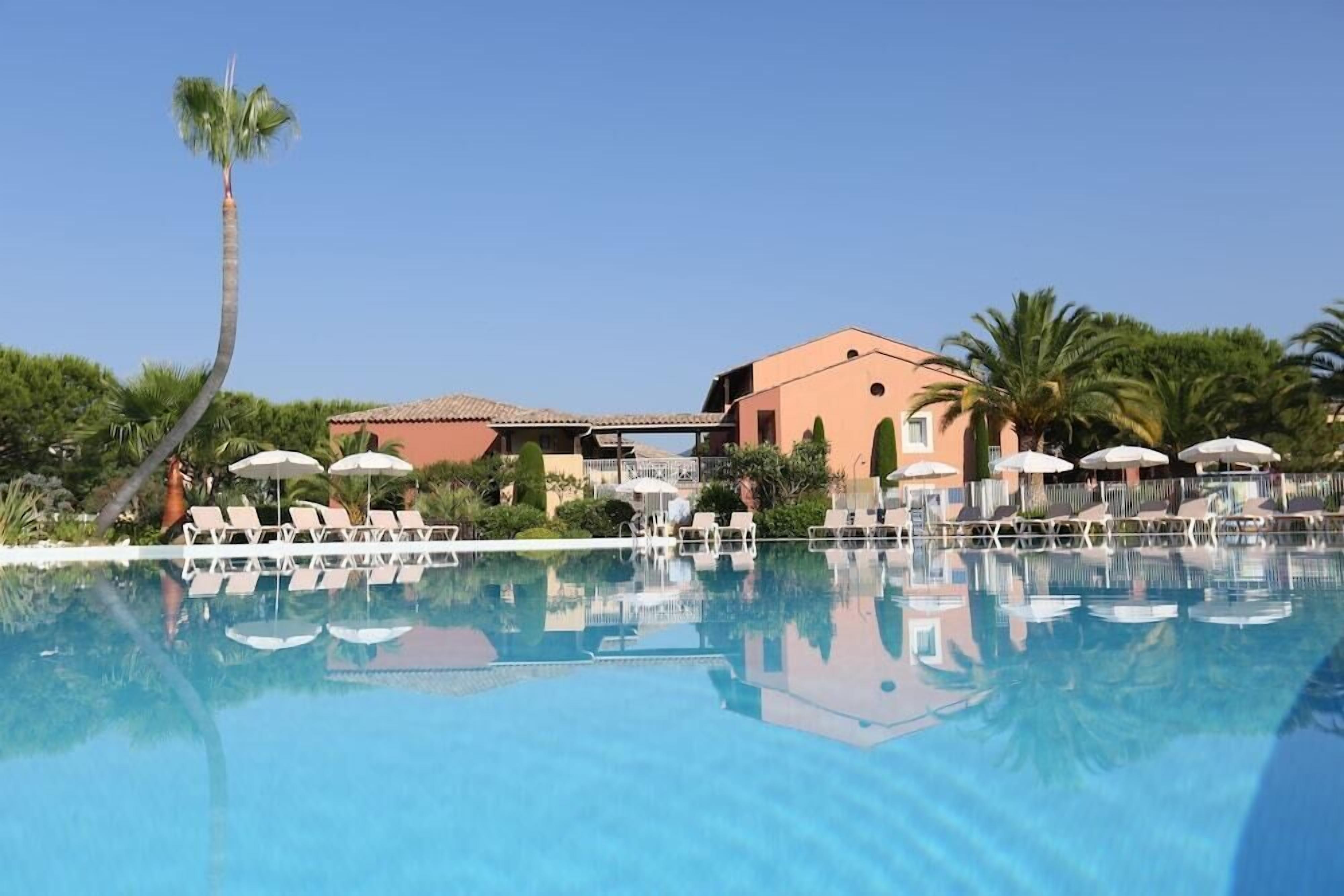 A clear blue pool with lounge chairs and umbrellas, surrounded by palm trees and a building with a tiled roof.